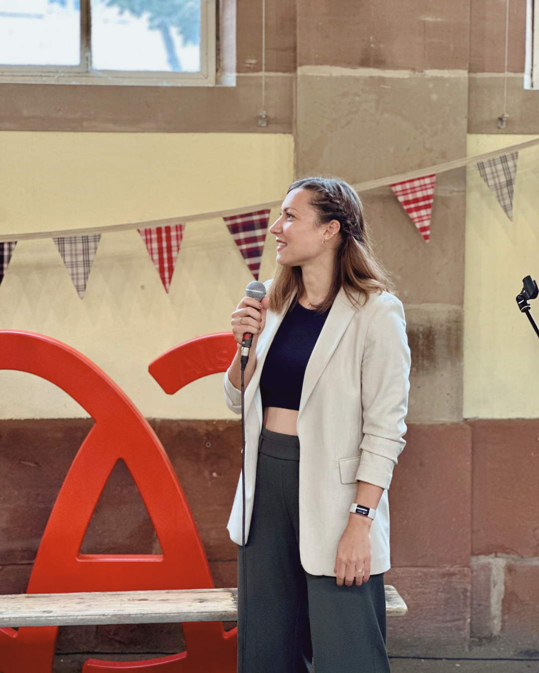 Tiffany, sophrologue à Strasbourg, tenant un micro lors d’une prise de parole en public, vêtue d’une veste claire et d’un top sombre, devant un décor coloré avec des fanions